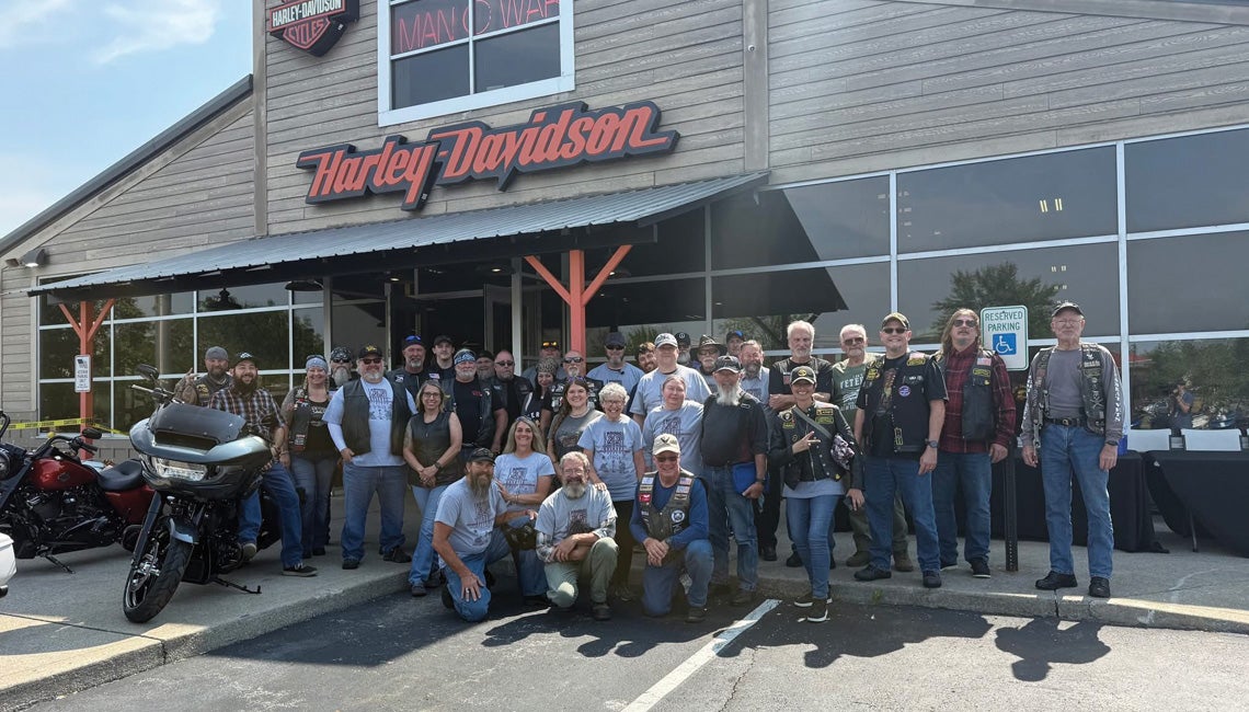 A large group of people posing with their motorcycles under a Harley-Davidson sign