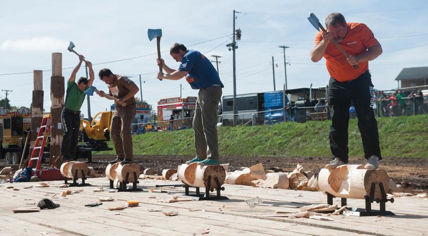 Four men swing axes while standing on wooden logs.