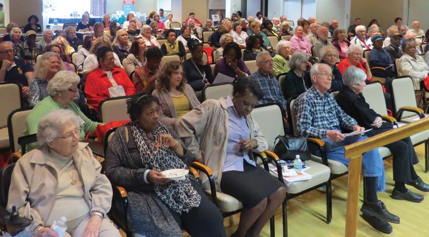 A large group of senior citizens sit and look toward a stage.