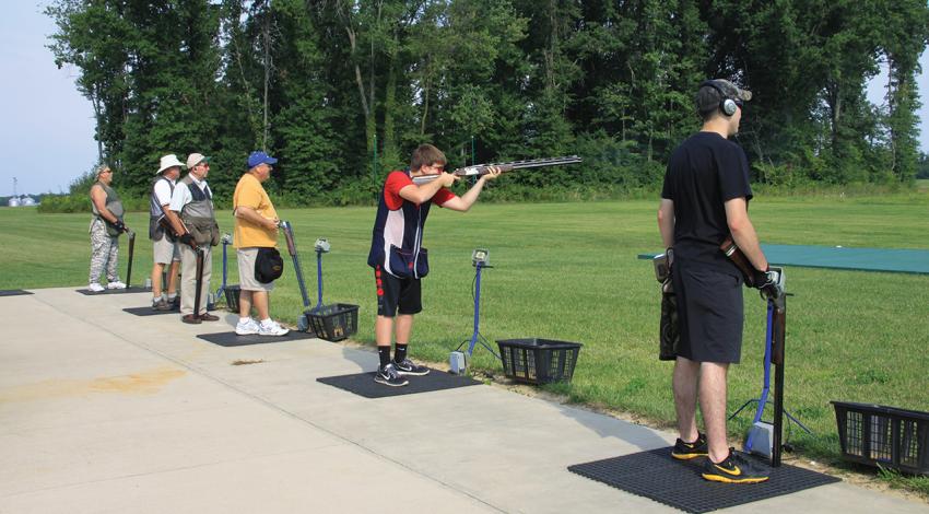 Six men stand at the shooting range, one pointing a gun.