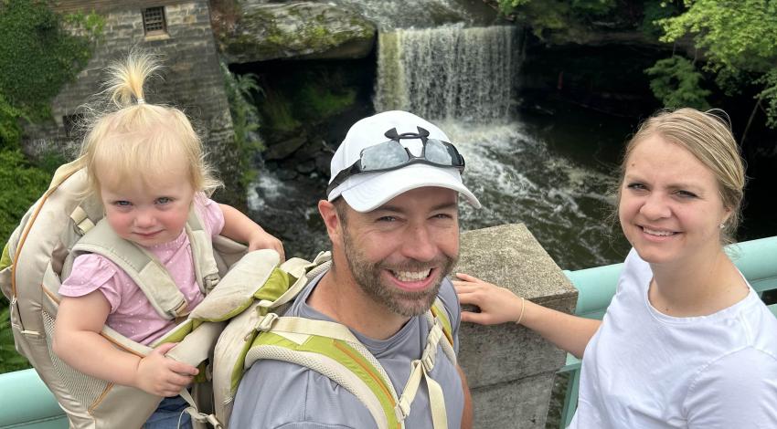 Man with baby in backpack and woman stand on bridge in front of a waterfall.