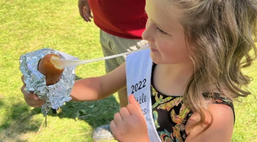 Little girl in crown and sash eating gooey fried cheese