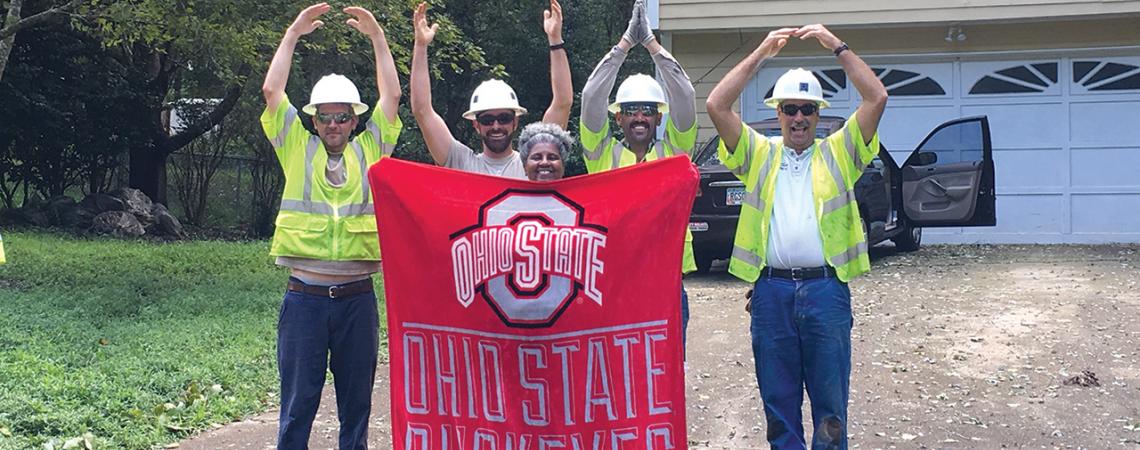 Linemen spell OHIO with their arms and surround a woman holding an Ohio State Buckeyes banner.