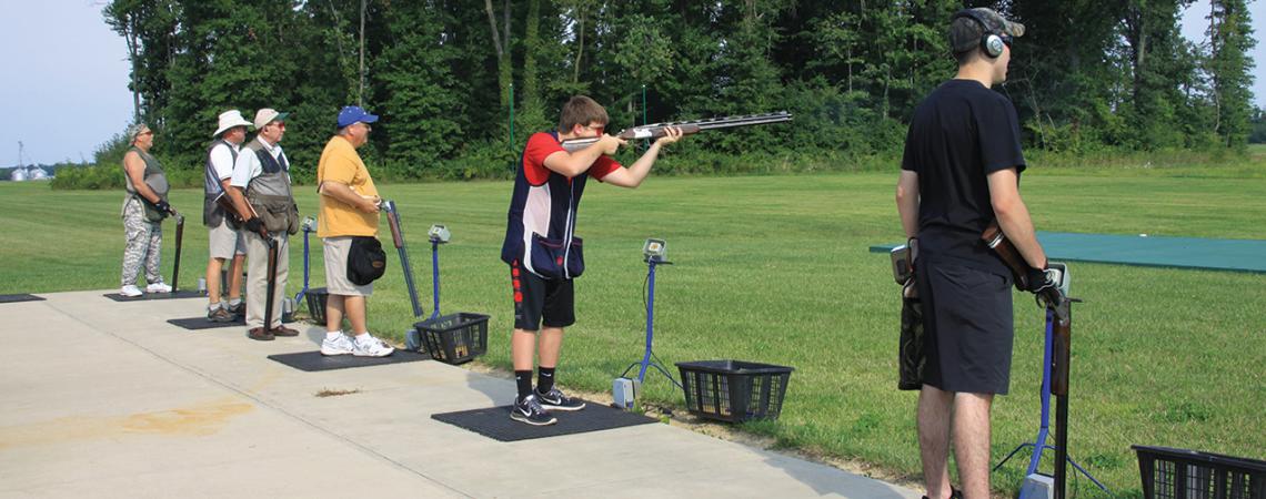 Six men stand at the shooting range, one pointing a gun.