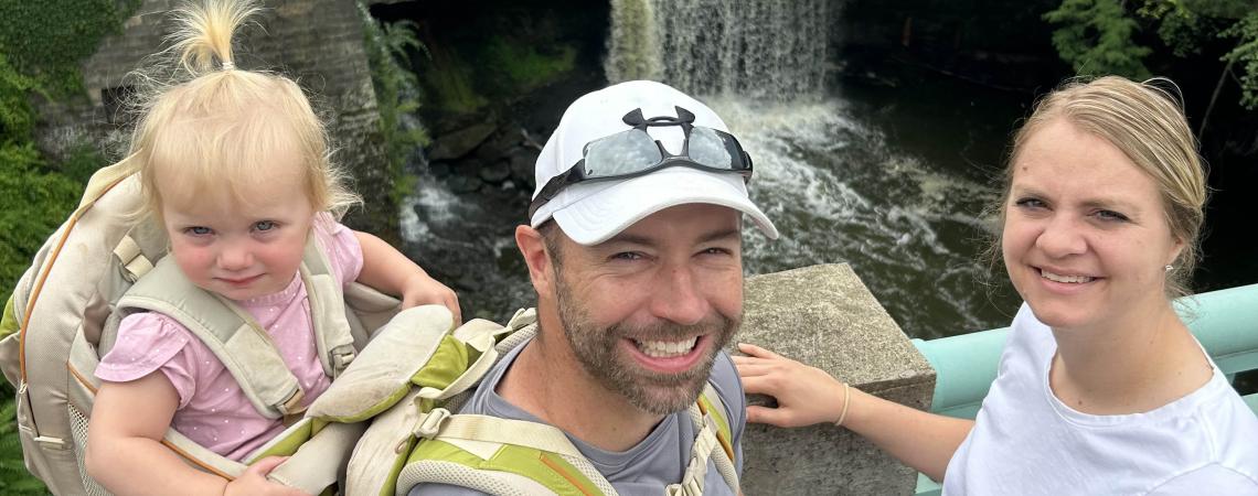 Man with baby in backpack and woman stand on bridge in front of a waterfall.